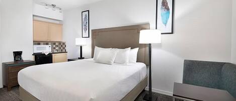 A hotel room with a large bed, a desk with a lamp, a chair, and a kitchenette in the background.
