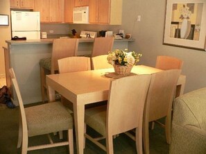 Un comedor con una mesa y sillas de madera, un cesto de flores y una cocina al fondo.