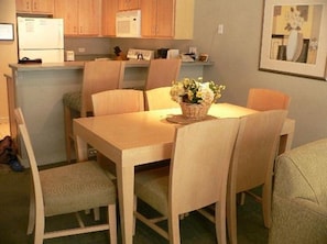 Un comedor con una mesa y sillas de madera, un cesto de flores y una cocina al fondo.