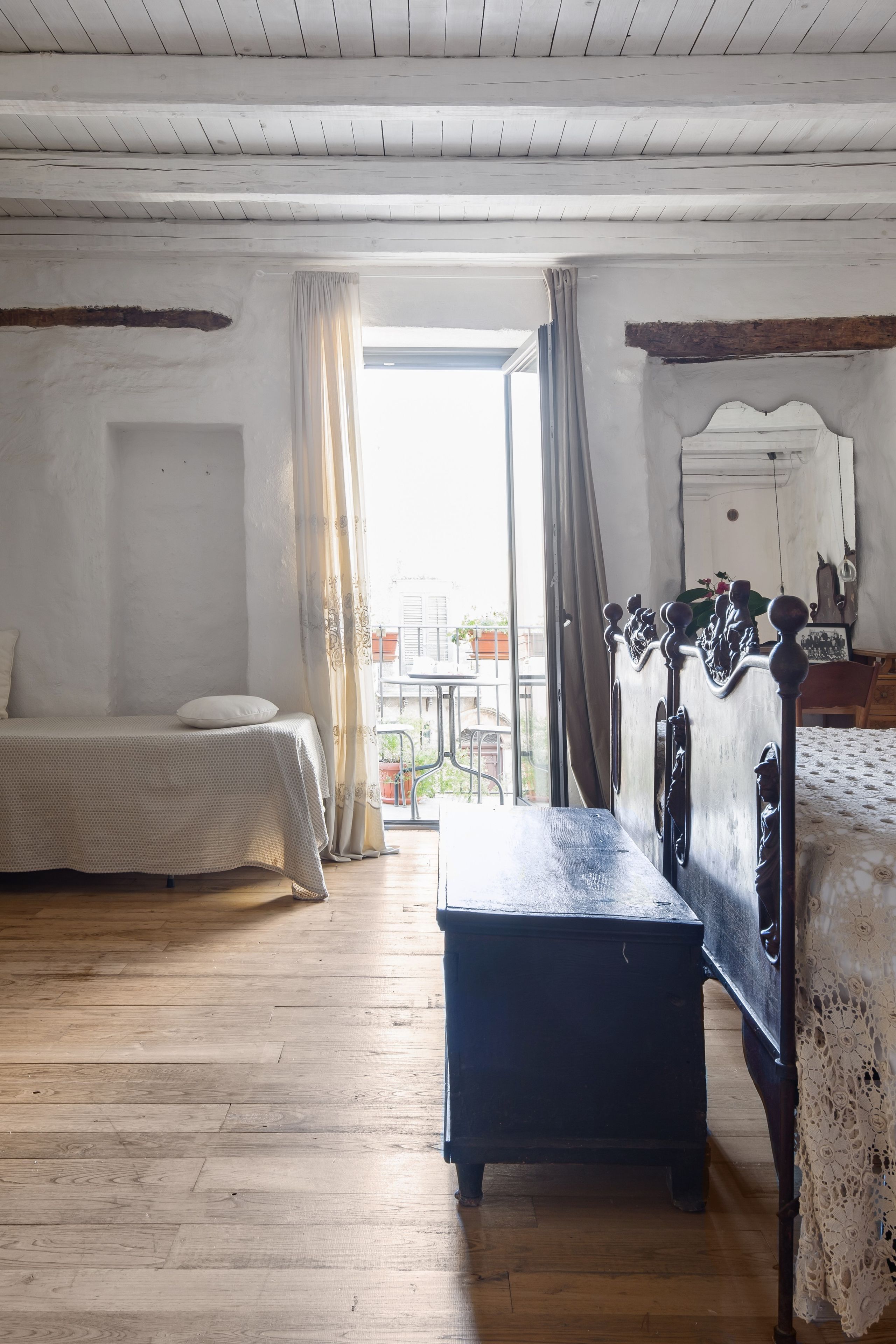 Ein Schlafzimmer mit Holzboden, einem Bett mit dunklem Kopfteil, einer Bank und einem Fenster mit Vorhängen.