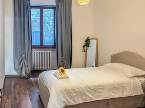 A bedroom with a bed, curtains, a radiator, a mirror, and a window with a view of stone walls.