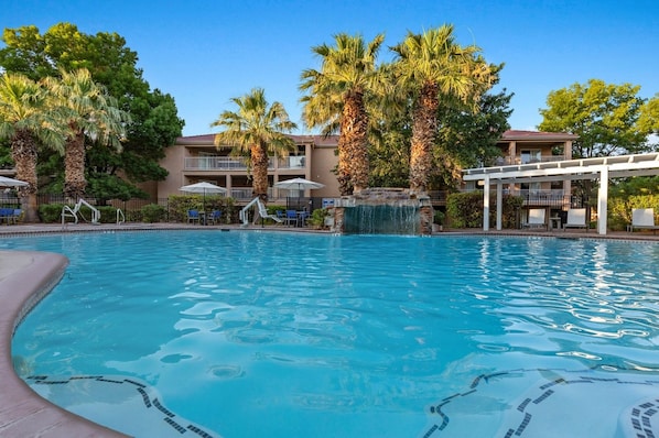 A swimming pool with palm trees and a waterfall feature, surrounded by lounge chairs and umbrellas. - St. George | Family Canyon Retreat | Pool + Hot Tub | 2BR (St. George)