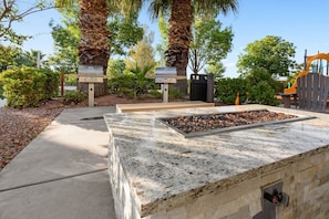 An outdoor fire feature with a stone base, surrounded by a concrete pathway and palm trees. - St. George | Family Canyon Retreat | Pool + Hot Tub | 2BR (St. George)
