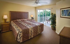 Habitación de hotel con una cama grande, una mesita de noche con lámpara, un ventilador de techo, un balcón con sillas y una planta en maceta.