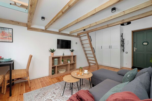 Ein gemütliches Wohnzimmer mit Holzbalkendecke, einem an der Wand montierten Fernseher, einem Sofa, einem Couchtisch, einem Korbstuhl und einer an der Wand angelehnten Leiter.