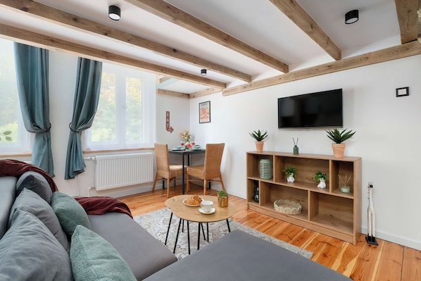 Un salón moderno con un sofá, una mesa de comedor con sillas, un televisor montado en la pared y una estantería de madera con plantas.
