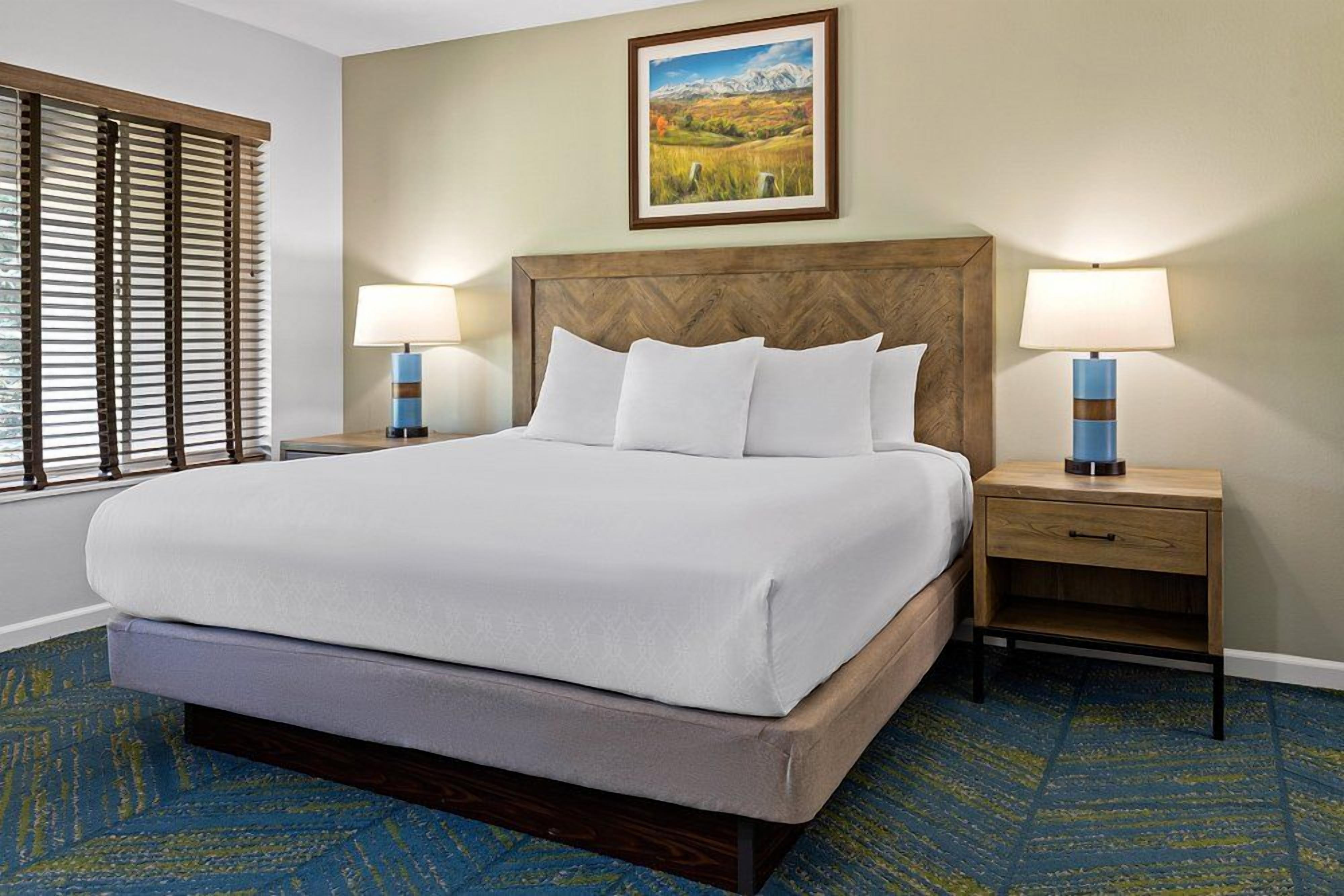 A hotel room with a large bed, two bedside tables with lamps, a painting on the wall, and a window with blinds.