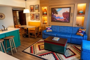 A living room with a blue sofa, a coffee table, a dining area with a table and chairs, and a kitchen area with a sink and stools. - Orlando | Family Lodge Retreat | Pool + Hot Tub | 2BR (Lake Buena Vista)