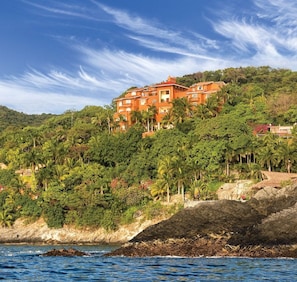Un hotel frente al mar con varios pisos, rodeado de exuberante vegetación y un litoral rocoso.