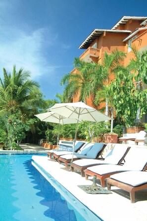 Una piscina con sillas de descanso y sombrillas, rodeada de palmeras y un edificio con balcón.