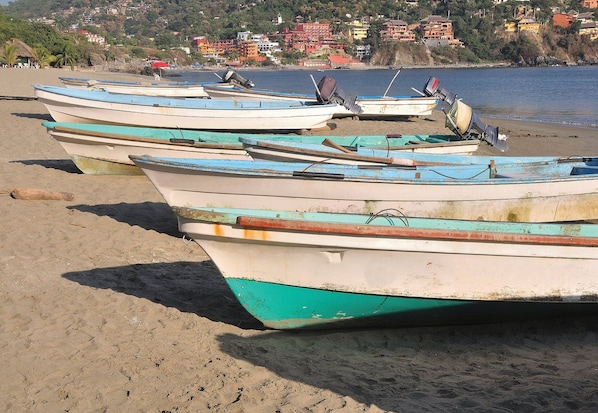 Varias embarcaciones alineadas en una playa arenosa, con un pueblo costero al fondo.