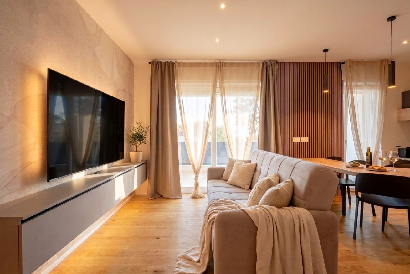 Ein modernes Wohnzimmer mit einem an der Wand befestigten Flachbildfernseher, einer Couch, einem Esstisch mit Stühlen und einem großen Fenster mit Vorhängen.