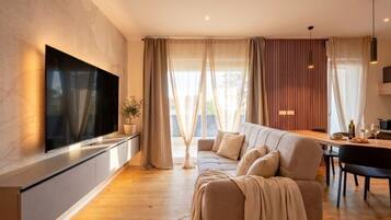 Ein modernes Wohnzimmer mit einem an der Wand befestigten Flachbildfernseher, einer Couch, einem Esstisch mit Stühlen und einem großen Fenster mit Vorhängen.