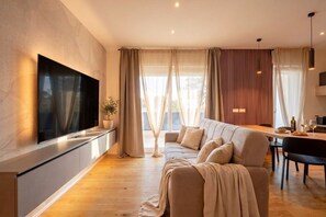Un soggiorno moderno con TV a schermo piatto montata a parete, un divano, un tavolo da pranzo con sedie e un'ampia finestra con tende.