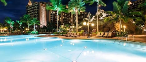 Une piscine nocturne, agrémentée de chaises longues et de palmiers, entourée d’immeubles dont les balcons sont illuminés.