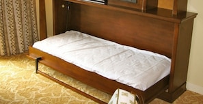 Un lit simple avec un cadre en bois et une literie blanche, dans une pièce dotée d’un tapis à motifs et d’un rideau à pois.