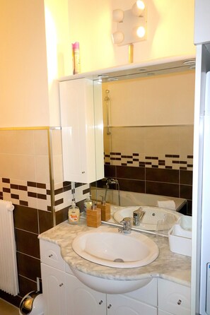 Et badeværelse med en hvid vask, et spejl og en bruser. Der er toiletartikler på bordet.