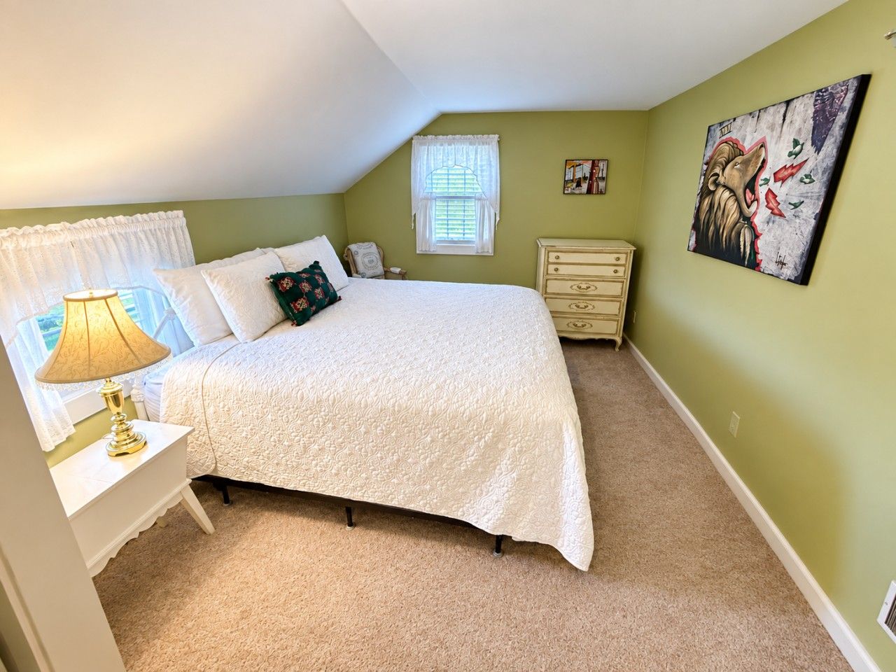 A bedroom with a bed, bedside table, lamp, window, dresser, and a painting on the wall.