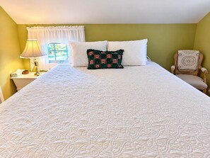 A bed with a quilted cover, a bedside lamp, a chair, and a window with curtains. - The Wiley | Cozy King Room with Bathtub (Peru)