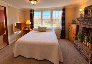 A bedroom with a large bed, a brick fireplace, a window with curtains, and a nightstand with a lamp. - The Wiley | Rustic Retreat with Jetted Tub and Corner Shower (Peru)