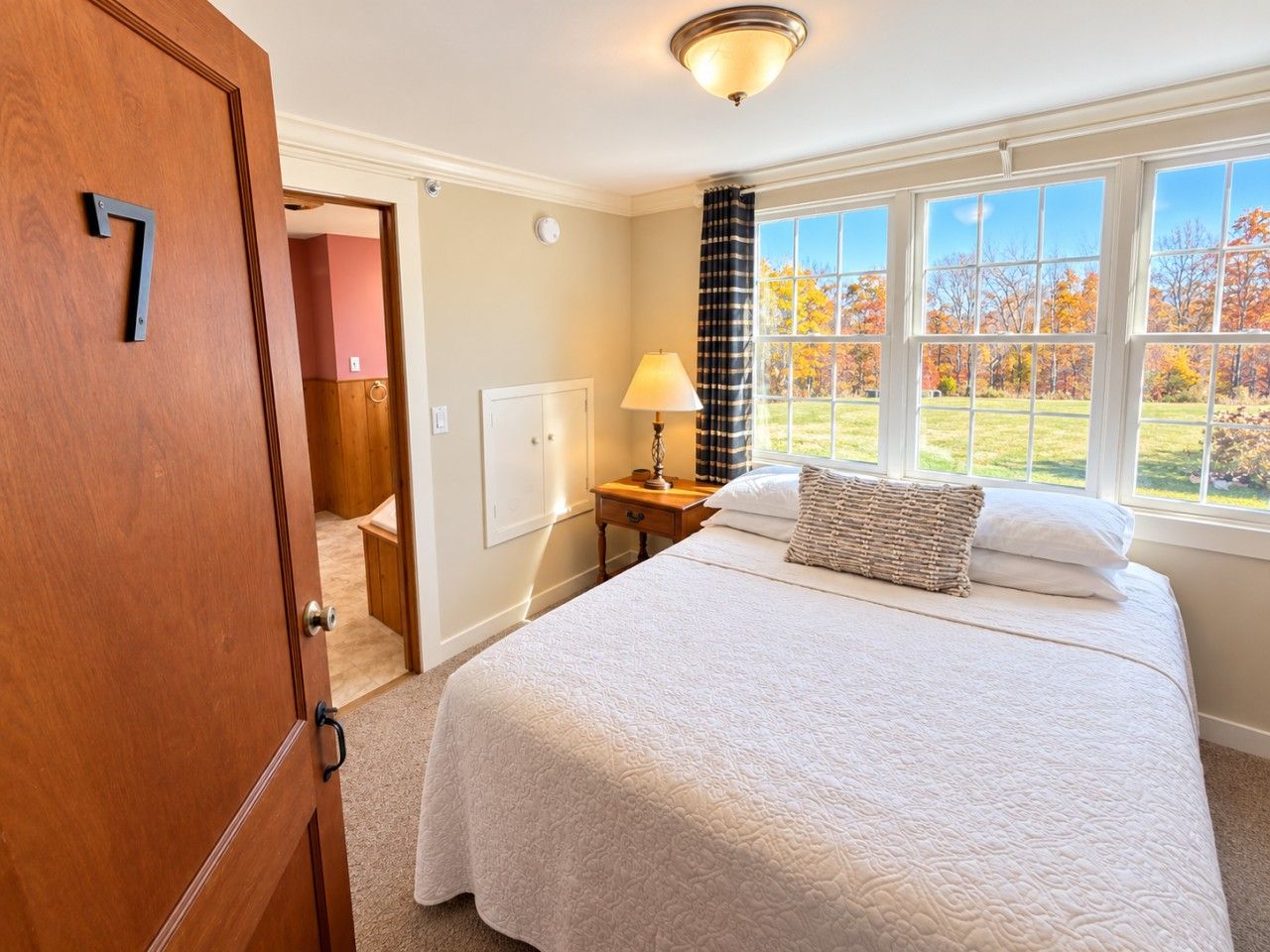 A bedroom with a bed, a nightstand, a lamp, a window with a view of trees, and a door with the number 7.