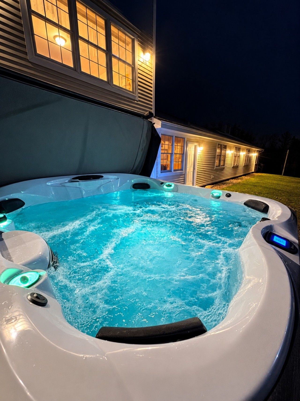 Un bain à remous doté d’un panneau de commande numérique, entouré d’une maison dont les fenêtres sont illuminées la nuit.