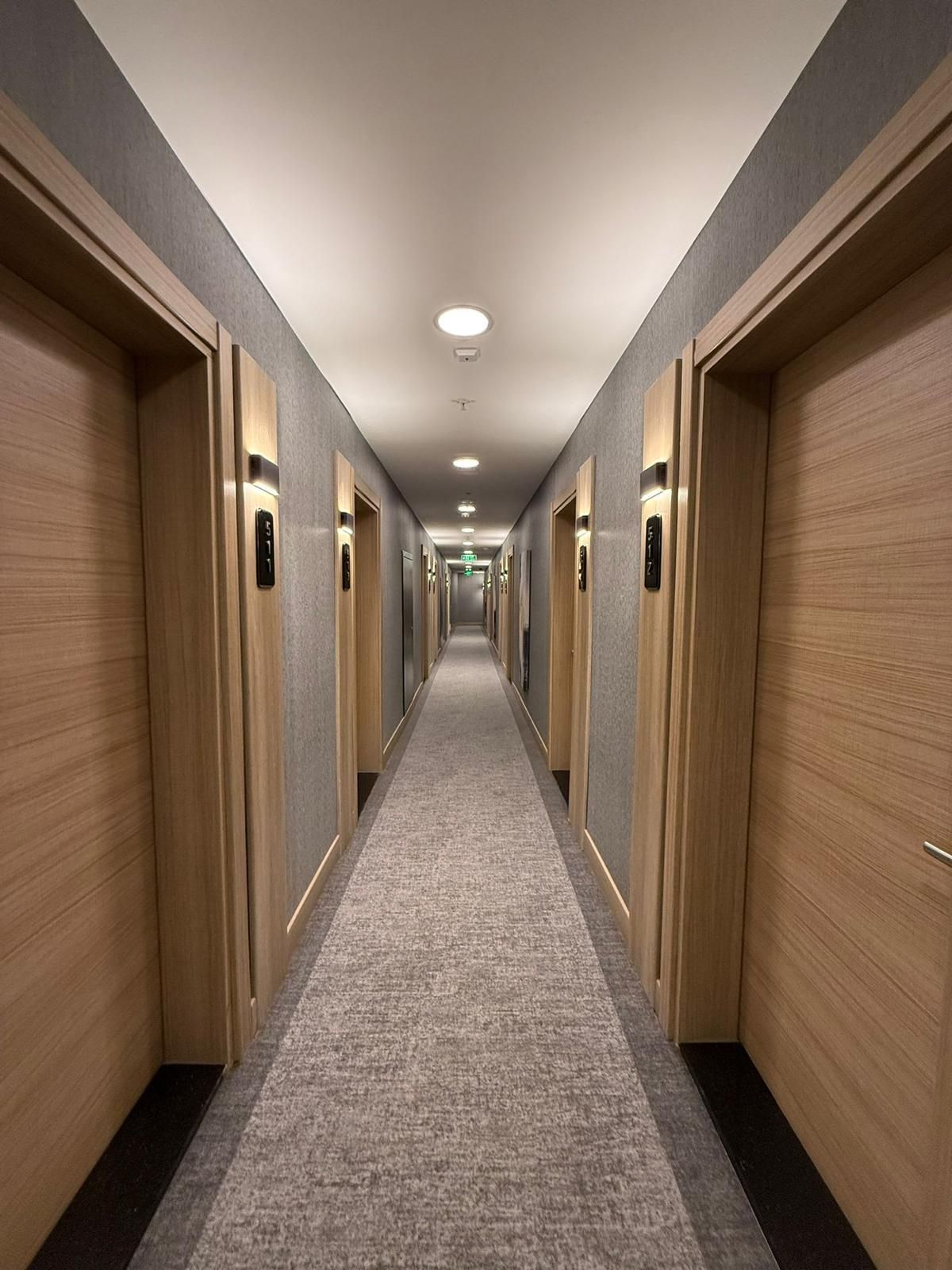 Un couloir d’hôtel avec des portes en bois, un sol recouvert de moquette et un éclairage encastré.