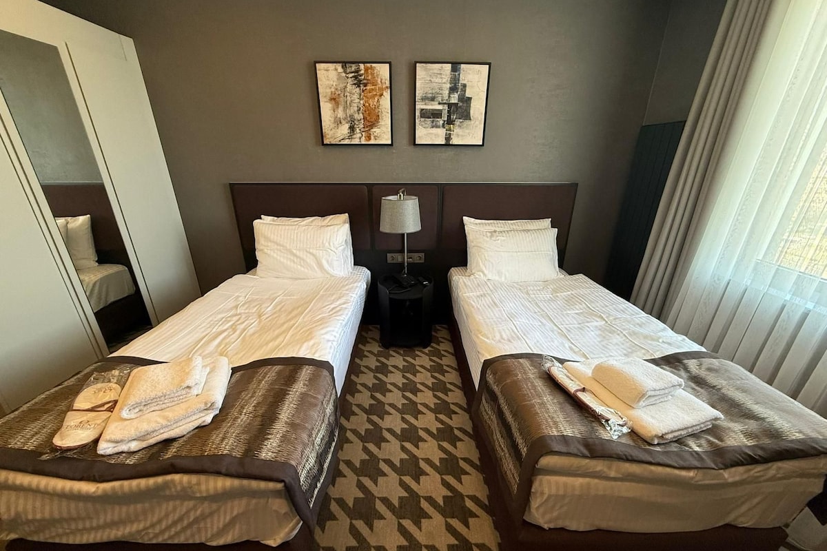 A hotel room with two single beds, a nightstand with a lamp, and two abstract paintings on the wall.