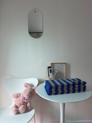 Une pièce avec une chaise blanche, un jouet en peluche rose, une petite table ronde sur laquelle sont pliées des serviettes, un tableau encadré et un miroir fixé au mur.