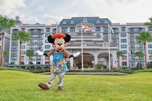 A person in a colorful costume standing in front of a hotel with multiple balconies and flags. - Orlando | Family Riviera Retreat | Pool + Resort | 2BR (Lake Buena Vista)
