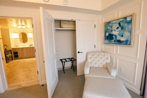 Ein Hotelzimmer mit Bett, Sessel, kleinem Tisch, Schrank mit Handtuchhalter und einem Bild an der Wand.