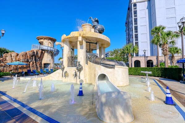 A water feature with a central fountain, multiple water jets, and a slide, surrounded by lounge chairs and palm trees. - Orlando • Grand Family Escape • Pool + Resort • 3BR Sleeps 12 (Lake Buena Vista)