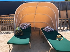 Una carpa en la playa con dos sillones reclinables verdes, cada uno con una toalla y un sombrero.