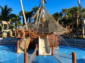 Tobogan d'aigua amb temàtica de vaixell pirata, una escala de corda, una piscina i palmeres al voltant.