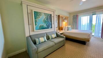 A hotel room with a sofa, a bed, a painting, a ceiling fan, and a window with curtains.