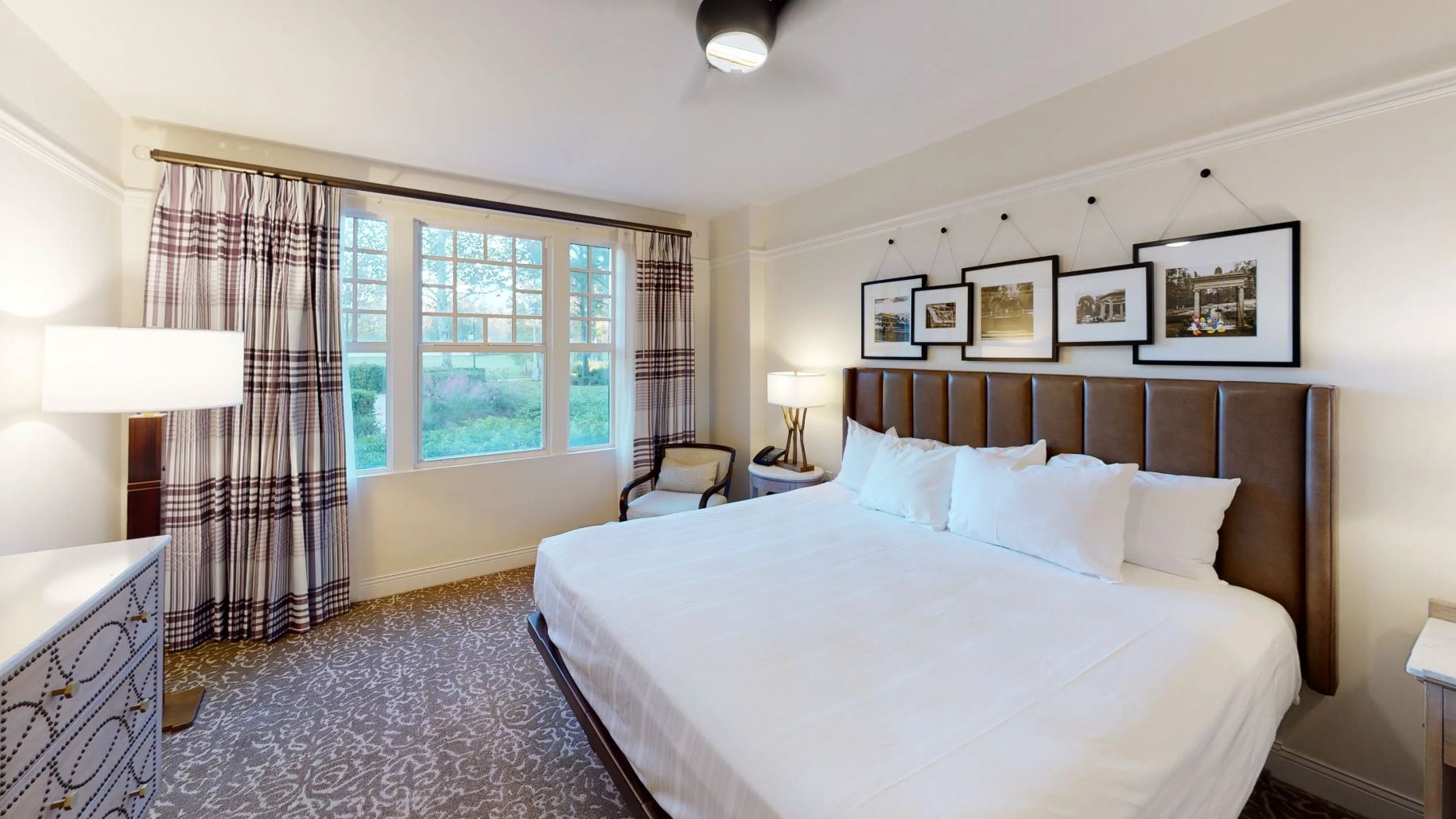 Quarto de hotel com uma cama grande, mesa de cabeceira, cadeira, janela com cortinas e quadros nas paredes.
