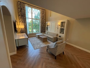 Een woonkamer met een bank, fauteuil, salontafel en een televisie. Er is een raam met gordijnen, een lamp en een kast met planken.