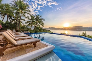 Una piscina infinita con sillas de descanso, palmeras y vistas al atardecer.