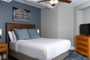 Cama bem arrumada com cabeceira de madeira, televisão sobre cômoda de madeira e ventilador de teto.