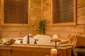 Interior de una cabaña de madera con una bañera, dos grifos, una vela y una toalla.