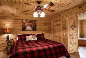 Un dormitorio de cabaña de madera con una cama, un ventilador de techo, un cuadro y una mesita de noche con una lámpara.