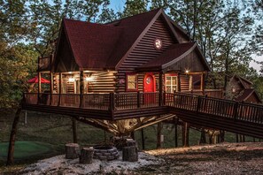 A wooden cabin with a red door, a balcony, and a staircase leading to the main entrance. - Branson | Family Treehouse Retreat | Pools + Fitness | 2BR (Ridgedale)