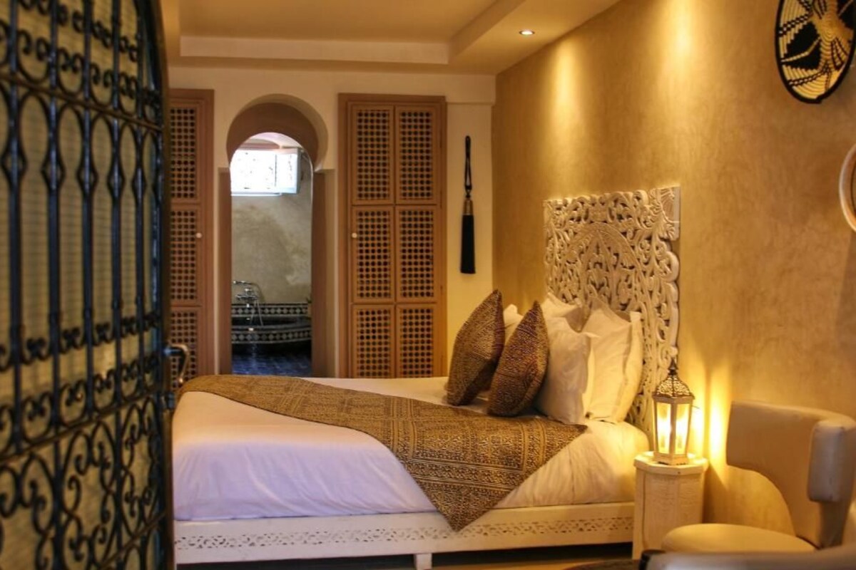 A hotel room with a bed, a nightstand, a lamp, and a view of a courtyard through an arched doorway.