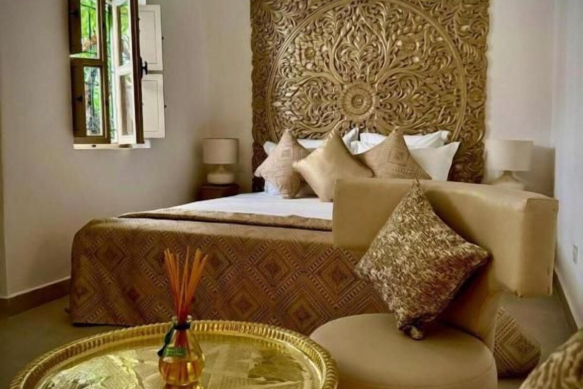 A room with a bed, a sofa, a round table with a decorative item, and a large ornate wall hanging.