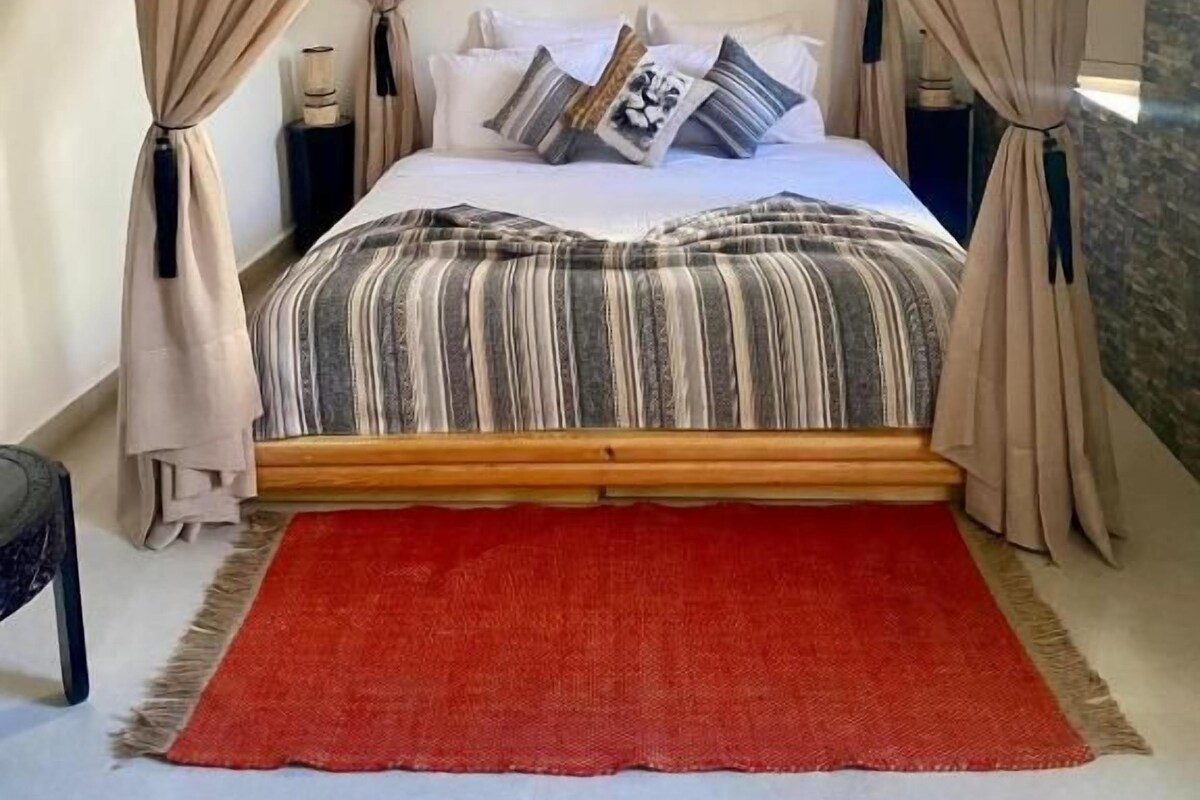 A four-poster canopy bed with a red rug, a striped bedspread, and a nightstand with a lamp.