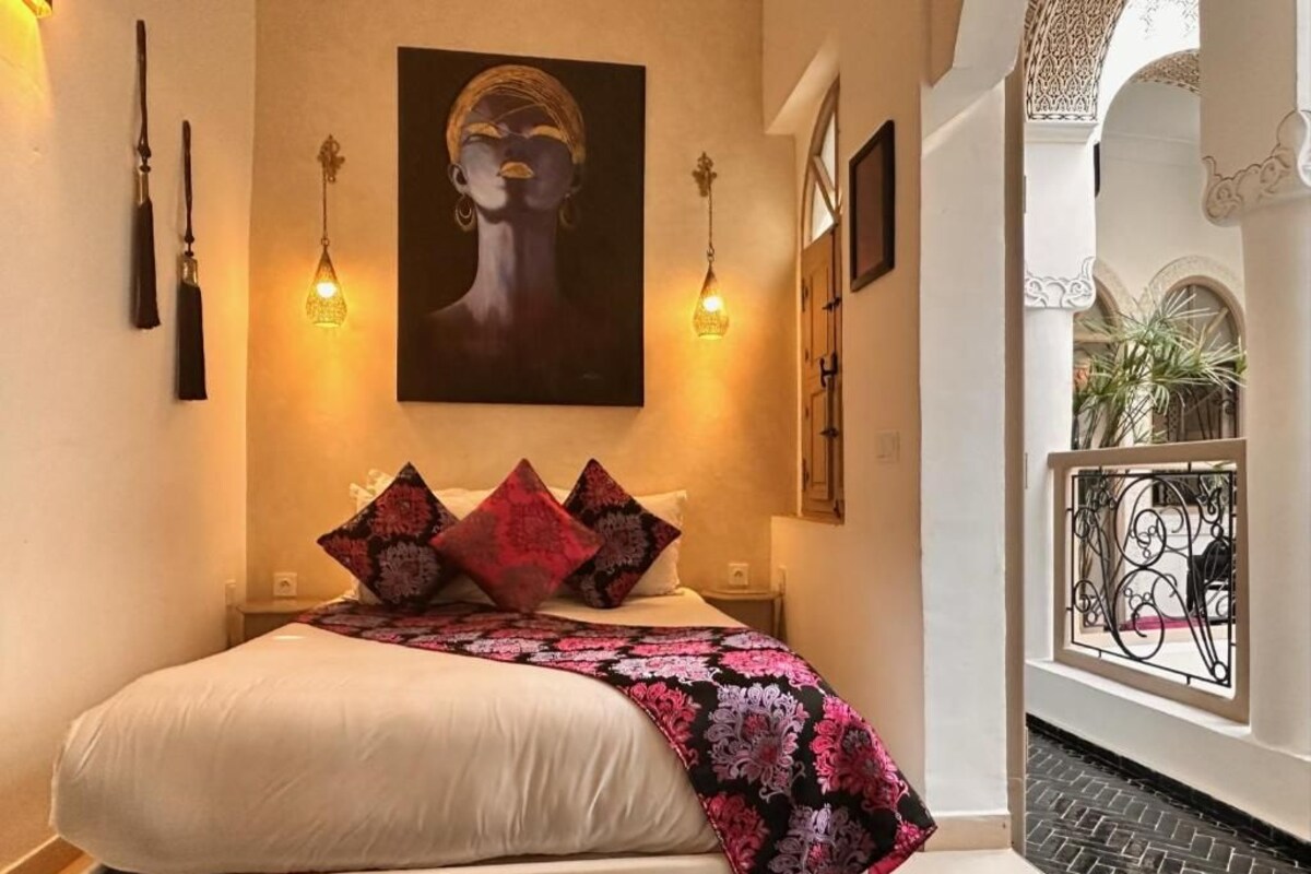 A bedroom with a bed, pillows, a blanket, a painting, and a balcony with a railing.