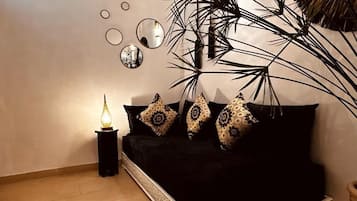 A room with a black sofa, decorative pillows, a small table with a lamp, and a large plant.
