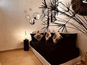 Una habitación con un sofá negro, almohadones decorativos, una mesita con una lámpara y una planta grande.