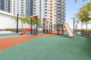 A playground with a slide and climbing structures, surrounded by a green turf area and a building in the background.