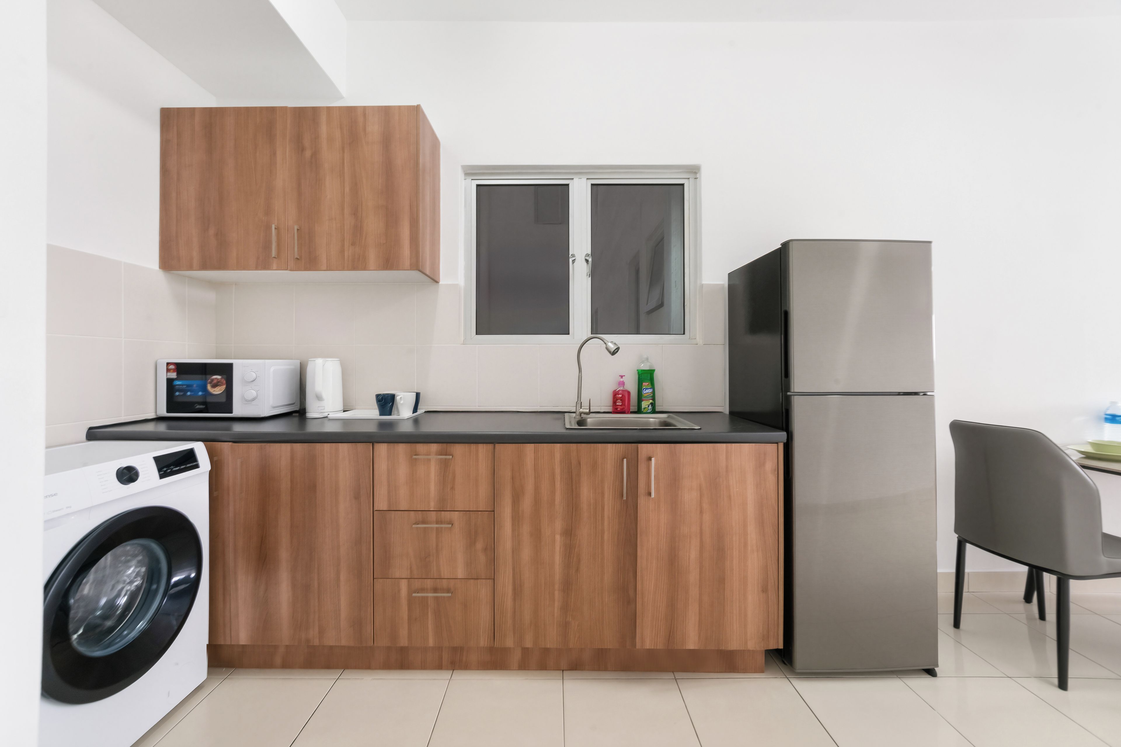 A modern kitchen with wooden cabinets, a stainless steel refrigerator, a washing machine, and a window above the sink.